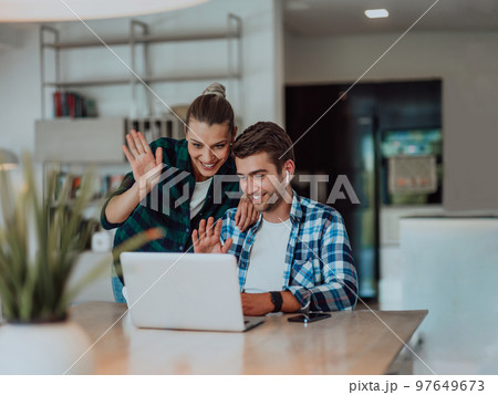 A young married couple is talking to parents, family and friends on a video call via a laptop while sitting in the living room of their modern house 97649673