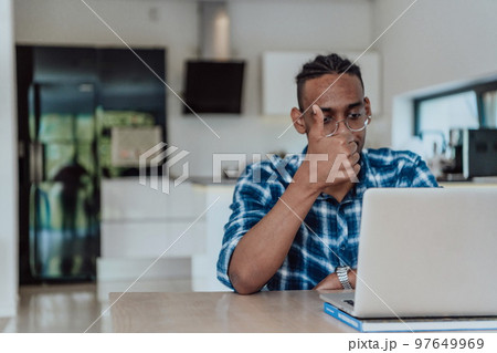 African American man in glasses sitting at a table in a modern living room, using a laptop for business video chat, conversation with friends and entertainment African American man in glasses sitting at a table in a modern living room, using a laptop for business video chat, conversation with friends and entertainment 97649969