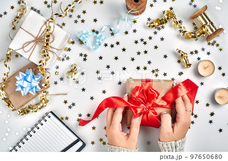Female hands holding present with red bow on white background with delicate wavy ribbons and metallic star shaped confetti. Flat lay style 97650680