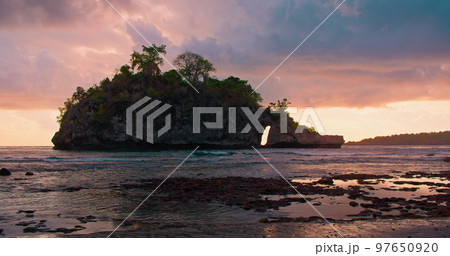 Beautiful sunset over mountain island Crystal Bay Beach Nusa Penida on background rear view of lonely woman in swimsuit walking along ocean waves. Orange light reflected on water surface. 97650920