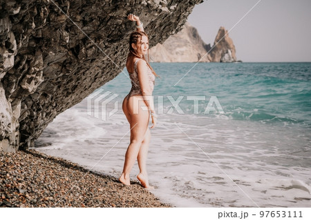 A tourist woman in bikini on beach enjoys the turquoise sea during her summer holiday. Beach vacation. Woman with braids dreadlocks standing with her arms raised near basalt rock enjoying beach ocean A tourist woman in bikini on beach enjoys the turquoise sea during her summer holiday. Beach vacation. Woman with braids dreadlocks standing with her arms raised near basalt rock enjoying beach ocean 97653111