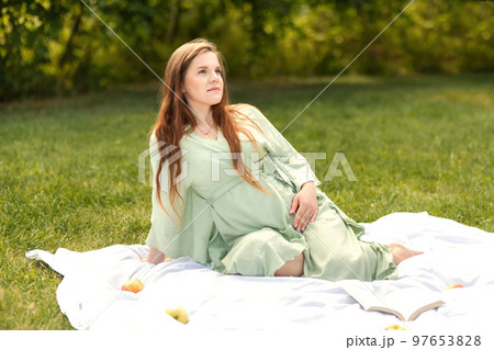 Young pretty pregnant woman resting in park, lying on cover,blanket in park,meadow, enjoying pregnancy, apples and book are beside. Healthy walk outdoors. Preparation for childbirth. Horizontal 97653828