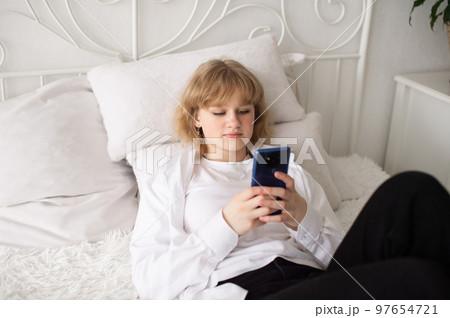 A cute girl in a white shirt and with a phone is lying on the bed. White interior 97654721