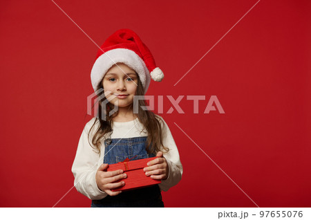 Lovely Caucasian little child girl in Santa hat, white pullover and blue denim overalls, smiling looking at camera, posing with cute gift box with surprise for Christmas, on red background. Ad space Lovely Caucasian little child girl in Santa hat, white pullover and blue denim overalls, smiling looking at camera, posing with cute gift box with surprise for Christmas, on red background. Ad space 97655076