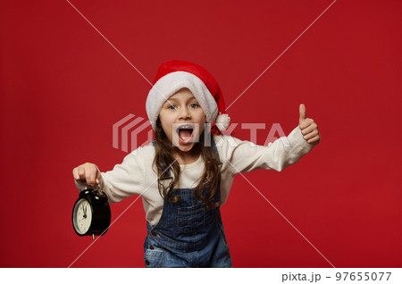 Overjoyed Caucasian 5 years old child girl with long hair, wearing Santa hat showing thumb up, posing with alarm clock showing midnight, looking at camera, red background. Happy New Year. Merry Xmas Overjoyed Caucasian 5 years old child girl with long hair, wearing Santa hat showing thumb up, posing with alarm clock showing midnight, looking at camera, red background. Happy New Year. Merry Xmas 97655077