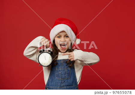 Overjoyed mischievous little girl in Santa hat, pointing at a vintage alarm clock with five to midnight, excited by the upcoming New Year event, isolated over red background. Copy advertising space 97655078