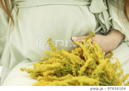 Cropped big belly of pregnant woman with yellow flowers Goldenrod.White female wears dress.Magic happy pregnancy. Childbirth preparations, emotional connection with baby. Baby shower. Horizontal 97656104
