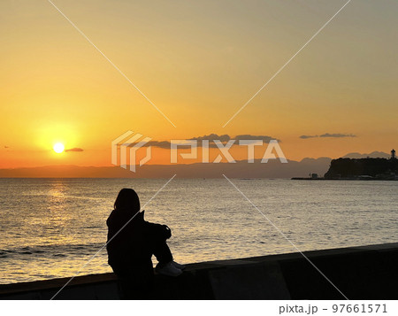夕焼けの海を膝を抱えて眺める女性のシルエットと江の島 97661571