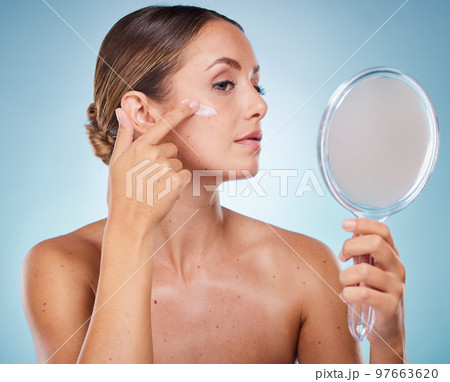 Woman, face and cream with hand mirror for beauty wellness, skincare dermatology and cosmetics moisturizer in blue background studio. Model, apply sunscreen and makeup facial product for self care 97663620
