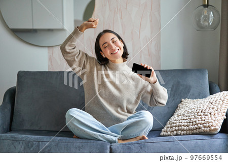 Happy and carefree girl singing and listening to music on smartphone app, using mobile phone as microphone, sitting on sofa 97669554