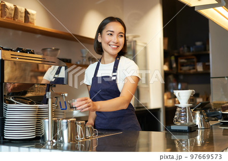 Smiling girl barista in cafe, preparing cappuccino in coffee machine, steaming milk, wearing uniform apron Smiling girl barista in cafe, preparing cappuccino in coffee machine, steaming milk, wearing uniform apron 97669573