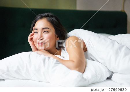 Dreamy asian girl lying in her bed in morning, covered with warm duver and leaning on white pillow, looking at window and thinking Dreamy asian girl lying in her bed in morning, covered with warm duver and leaning on white pillow, looking at window and thinking 97669629