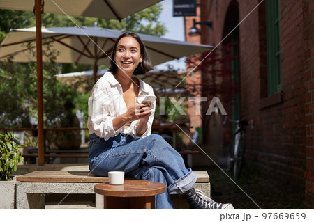 Stylish young asian woman sitting with cup of coffee in an outdoor cafe, smiling and looking happy, looking around Stylish young asian woman sitting with cup of coffee in an outdoor cafe, smiling and looking happy, looking around 97669659