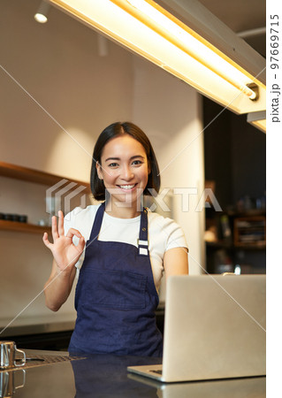 Portrait of barista girl, asian female cafe worker, shows okay, ok sign in approval, recommend smth, happy and pleased, wearing apron, standing with laptop behind counter 97669715