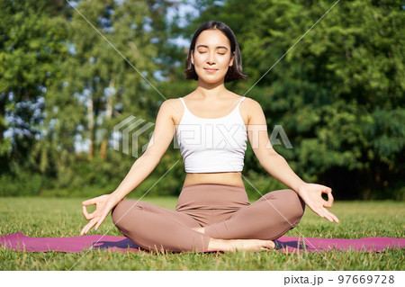 Sporty woman meditating on fresh air, sitting on fitness mat and practice yoga, smiling pleased. Sport and people concept 97669728