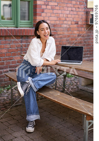 Vertical shot of good-looking asian woman with laptop, sitting in outdoor cafe, drinking coffee and smiling, laughing carefree Vertical shot of good-looking asian woman with laptop, sitting in outdoor cafe, drinking coffee and smiling, laughing carefree 97669766