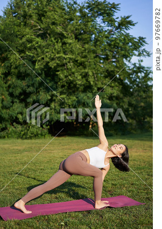 Vertical shot of young korean woman doing yoga training on rubber mat, making asana exercises on fresh air in park Vertical shot of young korean woman doing yoga training on rubber mat, making asana exercises on fresh air in park 97669782