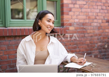 Beautiful young female model sitting with laptop in outdoor cafe, taking notes, writing in her diary or working on project, doing homework 97669791