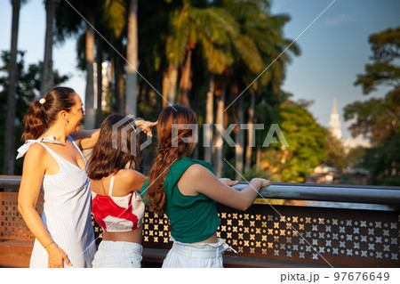 Tourists at one of the bridges along the Cali River Boulevard in the city of Cali in Colombia. Mother and teenager daughters traveling concept. Happy family concept. Tourists at one of the bridges along the Cali River Boulevard in the city of Cali in Colombia. Mother and teenager daughters traveling concept. Happy family concept. 97676649