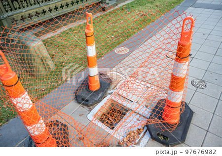 Open cover on a sidewalk access panel with hazard cones and fencing. 97676982
