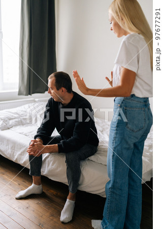 Vertical shot of despaired young husband sitting on bed looking away ignoring angry blonde wife arguing blaming upset man of problems at home. Jealous blonde woman shouting at sad boyfriend. 97677291