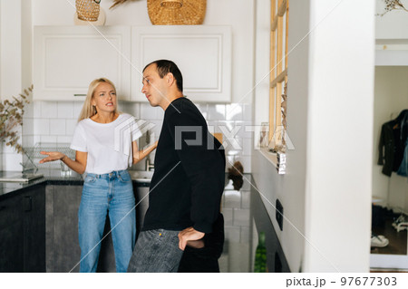 Angry wife scolding, raising voice, gesturing with hands, yelling at ignoring tired husband standing in kitchen room looking away. Concept of family scandal, crisis, domestic violence, abuse. 97677303