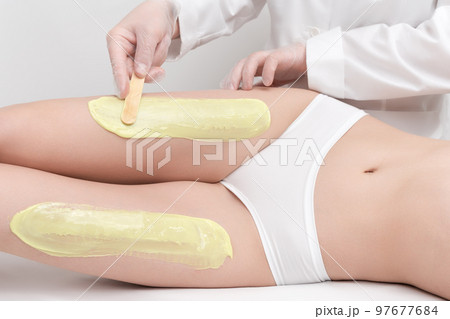 Beautician in gloves applying hot wax on slim woman legs using spatula while woman lying down on couch. Depilation with hot wax in beauty salon. Healthy body in white panties. Part of photo series. Beautician in gloves applying hot wax on slim woman legs using spatula while woman lying down on couch. Depilation with hot wax in beauty salon. Healthy body in white panties. Part of photo series. 97677684