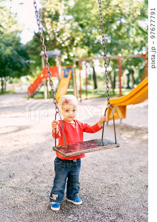 Little girl holding on to the handles of a wooden swing while standing on the playground. High quality photo 97677791