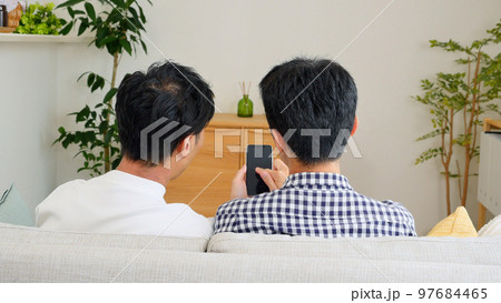 自宅でスマホを見て会話をする男性カップルの後ろ姿 97684465