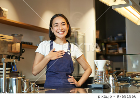 Friendly smiling asian girl barista, holds hands on heart, looks grateful at client, loves her guests in cafe, stands in uniform apron Friendly smiling asian girl barista, holds hands on heart, looks grateful at client, loves her guests in cafe, stands in uniform apron 97691966