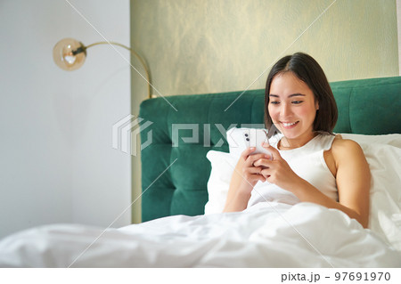 Beautiful smiling asian woman in bed, reading message on smartphone and laughing, enjoying lazy day in bedroom, using mobile phone 97691970