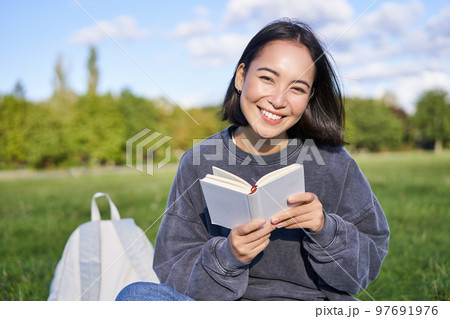 Beautiful woman sitting in park with book. Smiling asian girl reading and laughing, relaxing outdoors 97691976