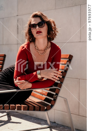 Portrait of a woman on the street. An attractive woman in glasses, a red blouse and a black skirt is sitting on a bench outside. Portrait of a woman on the street. An attractive woman in glasses, a red blouse and a black skirt is sitting on a bench outside. 97693977