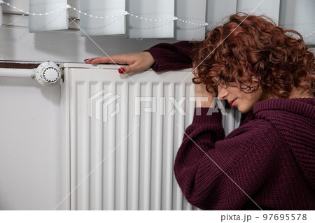 The girl, worried about the constant lack of heating, sits by the heater. 97695578