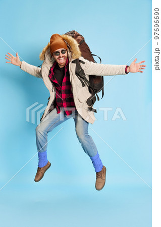Excited fashionable man in winter coat and hat with happy facial expression jumping, running isolated on blue background. Holidays, travel, adventure, ad 97696690