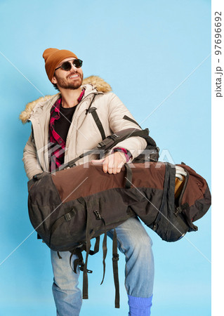 Delighted tourist. Happy man in winter coat and hat with joyful facial expression going to winter trip isolated on blue background. Holidays, travel, adventure, ad Delighted tourist. Happy man in winter coat and hat with joyful facial expression going to winter trip isolated on blue background. Holidays, travel, adventure, ad 97696692