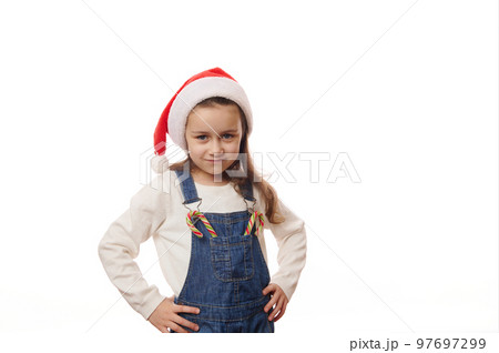 Adorable Caucasian little child girl wearing Santa hat, posing with hands on waist, with Christmas striped candy canes on the pocket of his denim overalls, looking at camera, against white background 97697299