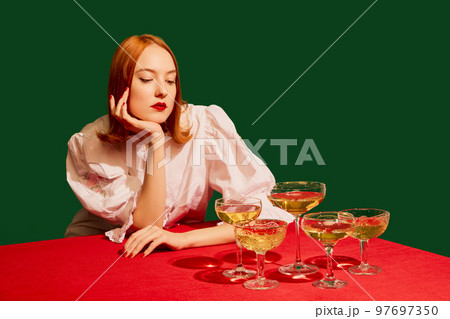 Young redhead girl in festive white dress sitting at the table with champagne over green background. Christmas mood 97697350
