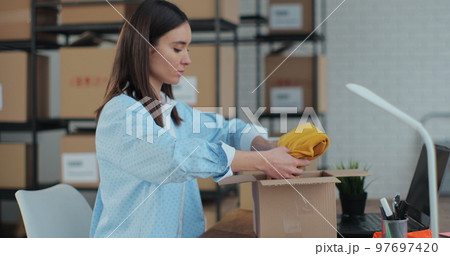 An employee packs a yellow jumper in a room with shelves full of parcels. The woman owner of an online store works on a computer at his desk in a warehouse. An employee packs a yellow jumper in a room with shelves full of parcels. The woman owner of an online store works on a computer at his desk in a warehouse. 97697420