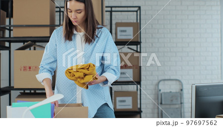 The owner of an online store works on a computer at her desk in a warehouse. An employee packs a yellow jumper in a room with shelves full of parcels. 97697562
