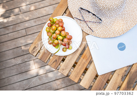 Closed laptop, grape, stylish hat, sunglasses are on a wooden table on a summer terrace with copy space. Summer mood, vacation, freelance, distance work concept. Top view. Background with copy space. 97703362