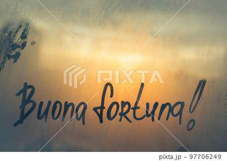 Lettering Italian text Buona fortuna Good luck in english message written finger on foggy glass wet sunset window 97706249