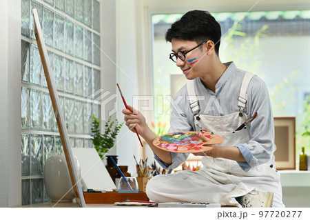 Smiling man artist sitting in front of canvas and painting picture with watercolor. Art and leisure activity concept 97720277