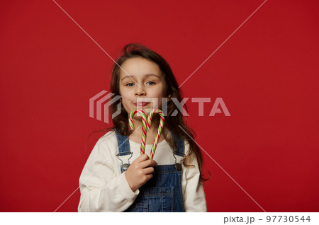 Beautiful little girl child holding sweet red striped green candy canes and cutely smiling looking at camera, isolated over red background with copy advertising space. Christmas and New Year concept Beautiful little girl child holding sweet red striped green candy canes and cutely smiling looking at camera, isolated over red background with copy advertising space. Christmas and New Year concept 97730544
