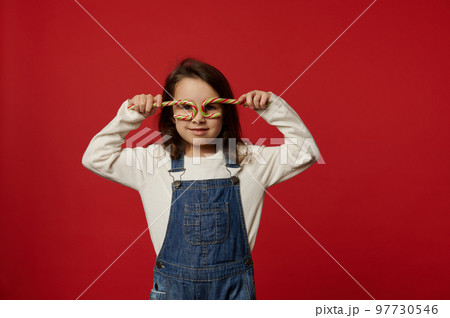 Adorable Caucasian baby girl wearing white pull and blue denim overalls, holding Christmas red and green striped lollipops candy canes like glasses, looking at camera, isolated on colored background 97730546