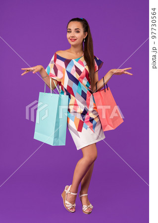 Beautiful young woman holding shopping bags and looking at the camera while standing against purple background. 97730564
