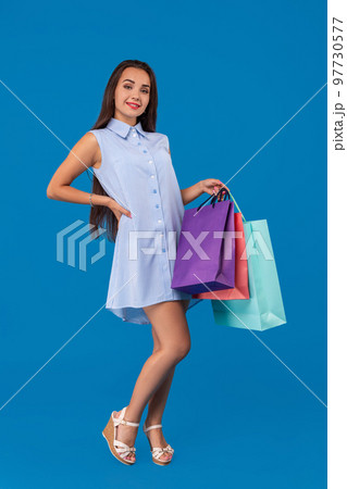 Beautiful young woman holding shopping bags and looking at the camera while standing against blue background. 97730577