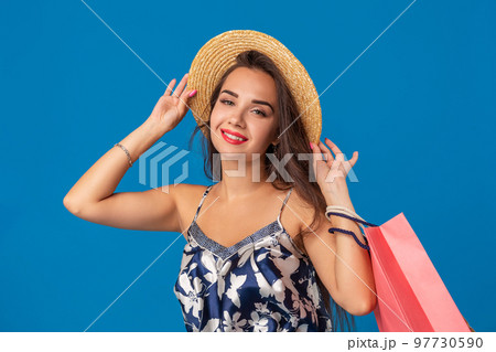 Portrait of a young woman in summer hat holding shopping bags and looking at the camera isolated over blue background 97730590