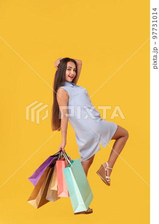 Image of a young brunette lady in white and blue summer striped dress posing with shopping bags and looking at camera over yellow background. 97731014