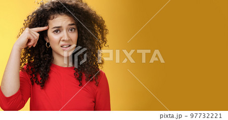Are you crazy. Portrait of frustrated and confused woman with curly hair holding index finger near temple and raising eyebrows shocked being annoyed with stupid actions of weirdo over yellow wall 97732221